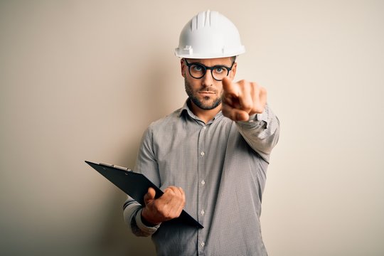 Young architect inspector man wearing builder safety helmet and using clipboard pointing with finger to the camera and to you, hand sign, positive and confident gesture from the front