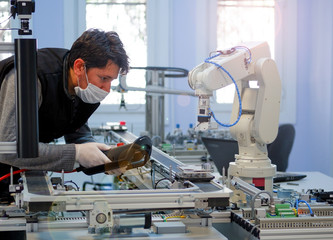 Man with surgical mask on face against COVID-19 ( Coronavirusis ) programming robot arm with control panel which is integrated on smart factory production line. industry 4.0 automation line.