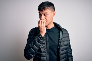 Young handsome hispanic man wearing winter coat standing over white isolated background looking stressed and nervous with hands on mouth biting nails. Anxiety problem.