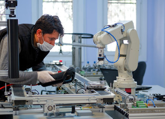 Man with surgical mask on face against COVID-19 ( Coronavirusis ) programming robot arm with control panel which is integrated on smart factory production line. industry 4.0 automation line.