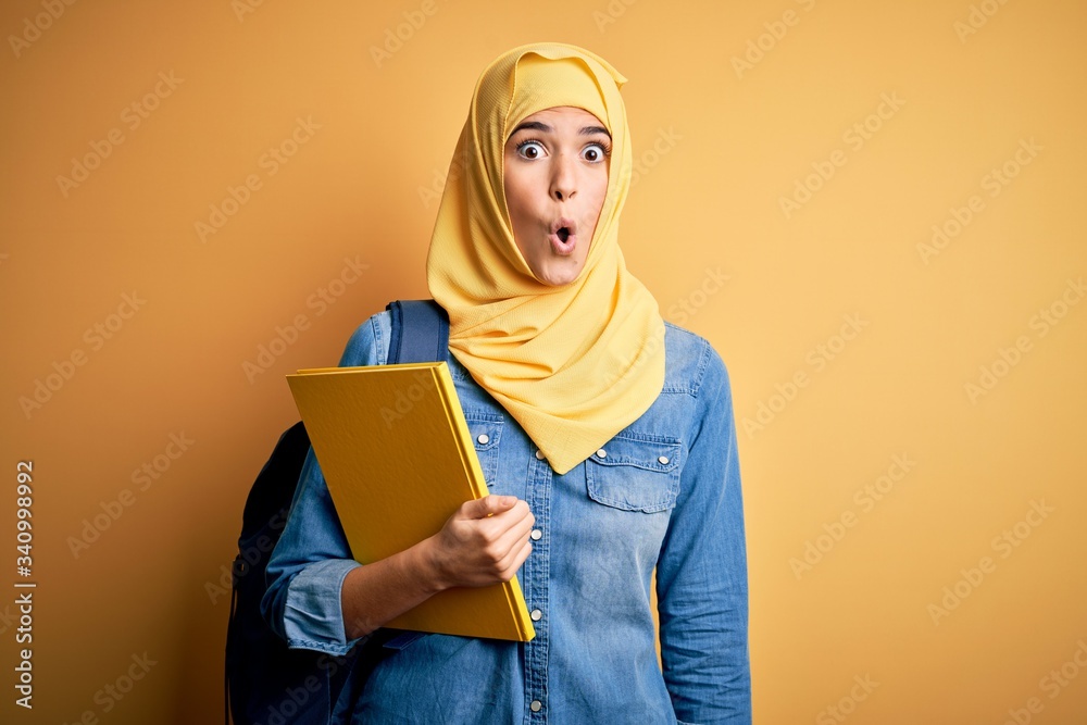 Sticker young student girl wearing muslim hijab and backpack holding book over yellow background scared in s