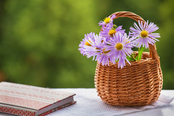 Naklejka na ściany i meble Lilac flowers in a wicker basket and a notebook are on the table. Blurred green background. Copy space.