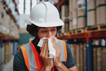 Young warehouse worker feeling sick while sneezing and coughing at factory standing near packed...