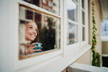 Beautiful young woman smiling drinking coffee and daydreaming enjoying outside view during isolation