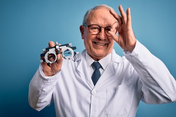Senior grey haired optic doctor man holding optometrist eyeglasses over blue background with happy face smiling doing ok sign with hand on eye looking through fingers