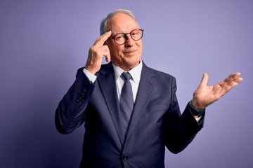 Grey haired senior business man wearing glasses and elegant suit and tie over purple background confused and annoyed with open palm showing copy space and pointing finger to forehead. Think about it.