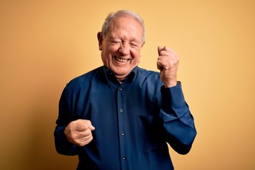 Grey haired senior man wearing casual blue shirt standing over yellow background celebrating surprised and amazed for success with arms raised and eyes closed. Winner concept.