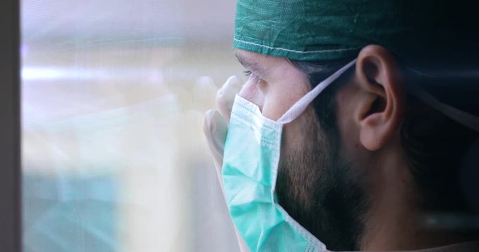 Medico o infermiere uomo con cuffia e mascherina guarda stanco fuori dalla finestra.