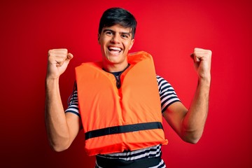 Young handsome man wearing orange safety life jacket over isolated red background celebrating...