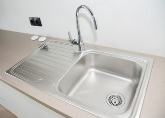 Kitchen countertop with a top mounted modern metallic kitchen sink and stainless steel water tap and black European electrical outlet mounted on the white wall.