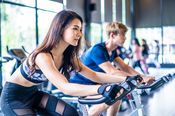Fototapeta na wymiar Young fit muscle Asian woman and Caucasian man exercising and working out in gym. People in sportswear cycling on exercise bike for cardio and focus on training for good health, wellness in fitness.