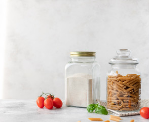 glass jars with whole grain flour and whole grain penne pasta. gluten free. smart food storage. copy space. Gray background.