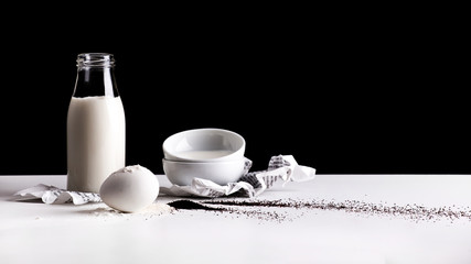 Breakfast composition made of milk, flour, egg, poppy seeds, napkins, little bowls on a white table with the black background