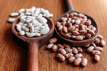 White and pinto beans on wooden kitchen spoons. Brown, wooden background.