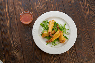 Fried spring rolls with shrimp, fresh arugula salad and sweet chili sauce, served in white plate over wooden texture background. Top view flat lay, copy space asian food concept