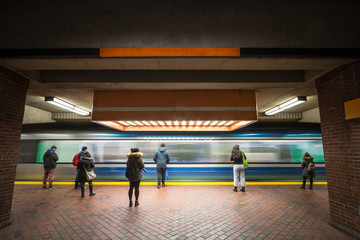 Metro entering fast in a peak transit station with passengers seen from behing standing immobile in...