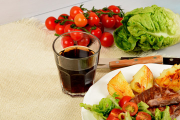 pieces of grilled steak next to potatoes grilled next to lettuce with tomatoes and olives and a drink in a glass