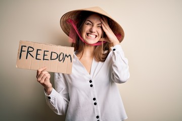 Young redhead woman wearing asian traditional hat holding banner with freedom message stressed with hand on head, shocked with shame and surprise face, angry and frustrated. Fear and upset