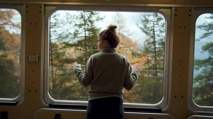 Cinematic and symmetrical beautiful shot of female traveler, travel blogger and inspired adventurer hang out of train window, look at amazing landscape of autumn mountains