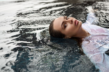 Portrait of mysterious sexy seductive woman in swimming pool water. Pretty alluring young girl wearing wet white shirt.
