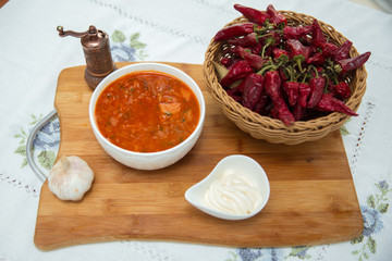 Garlic . A bowl of white ceramic salt and pepper. Ceramic plate Mayonnaise . Parsley Bowl of borscht soup on wooden board . Parsley . Red Dry Pepper in Traditional Basket on the tablecloth Table .