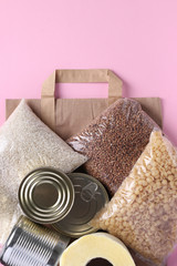 Paper bag with food supplies crisis food stock for quarantine isolation period on pink background. Rice, buckwheat, pasta, canned food, toilet paper. Food delivery, Donation, Vertical format, Top view