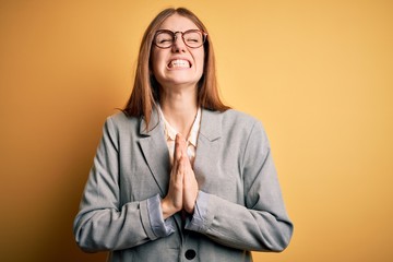 Young beautiful redhead woman wearing jacket and glasses over isolated yellow background begging...