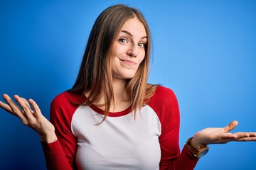 Young beautiful redhead woman wearing casual t-shirt over isolated blue background clueless and...