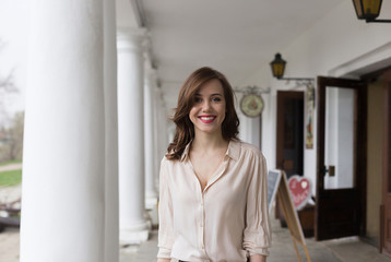 pretty caucasian young smiling girl in beige blouse with golden cross on her chest, seductive red lipstick and wavy hair standing on veranda of cafe. white concrete pillars and lanterns in background