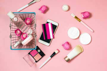 Obraz na płótnie Canvas Concept of online shopping cosmetics. Top view on cosmetics bottles, cream, soap, makeup brushes, mobile phone, credit card and shopping basket from the supermarket on a pink background, flat lay