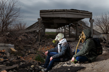Two people in gas masks sitting among ruins and dramatic sky background. Enviromental pollution, ecology disaster, radiation, biological hazard concept.