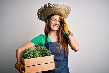 Young beautiful redhead farmer woman wearing apron and hat holding box with plants stressed with hand on head, shocked with shame and surprise face, angry and frustrated. Fear and upset for mistake.