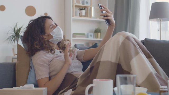 Lockdown of Caucasian curly-haired woman wearing medical mask sitting on couch in the apartment with smartphone in her hands and making selfie