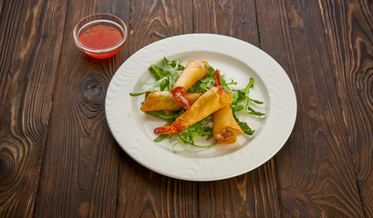 Fried spring rolls with shrimp, fresh arugula salad and sweet chili sauce, served in white plate over wooden texture background. Top view flat lay, copy space asian food concept
