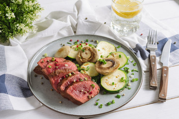 Rare roast beef with grilled zucchini and mushrooms. A glass of water with a slice of lemon. Utensils with wooden handles. Kitchen towel. Sprinkled with a mixture of peppers and chopped scallion.