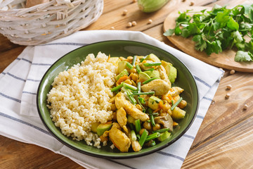 Green plate with chopped chicken with bulgur on the side dish, chickpeas and oyster mushrooms. The dish is sprinkled with chopped scallion. On a wooden kitchen table with a striped kitchen towel.