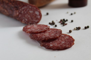 a stick of smoked raw sausage and slices cut into rings on a white background with peppers peas and baguette and a salt shaker in the background