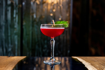 A red sour cocktail in a coupe glass, decorated with a fir branch on the table in a bar - obrazy, fototapety, plakaty