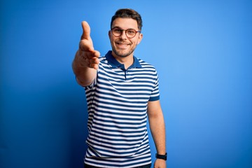 Young man with blue eyes wearing glasses and casual striped t-shirt over blue background smiling friendly offering handshake as greeting and welcoming. Successful business.