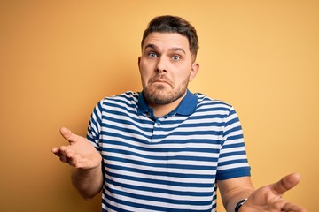 Young man with blue eyes wearing casual striped t-shirt over yellow background clueless and confused expression with arms and hands raised. Doubt concept.
