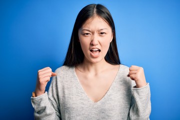 Young beautiful asian woman wearing casual sweater standing over blue isolated background angry and mad raising fists frustrated and furious while shouting with anger. Rage and aggressive concept.