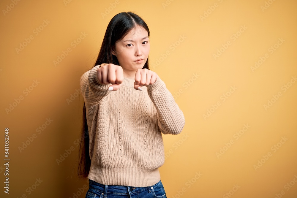 Poster Young beautiful asian woman wearing casual sweater over yellow isolated background Punching fist to fight, aggressive and angry attack, threat and violence