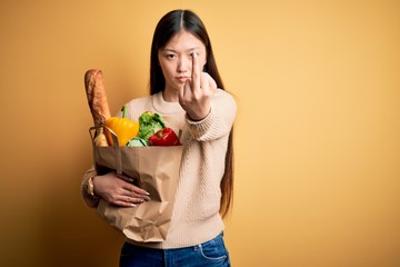 Young asian woman holding paper bag of fresh healthy groceries over yellow isolated background Showing middle finger, impolite and rude fuck off expression