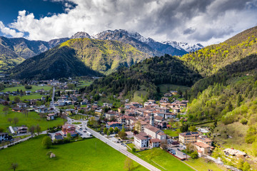 Fototapeta na wymiar Aerial view of improbable green meadows of Italian Alps, Comano Terme, huge clouds over a valley, roof tops of houses, Dolomites on background, sunshines through clouds