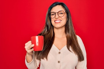 Young hispanic woman wearing glasses drinking a cup of coffee over red background with a happy face standing and smiling with a confident smile showing teeth