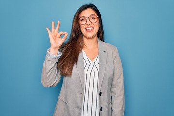 Young hispanic business woman wearing glasses standing over blue isolated background smiling positive doing ok sign with hand and fingers. Successful expression.