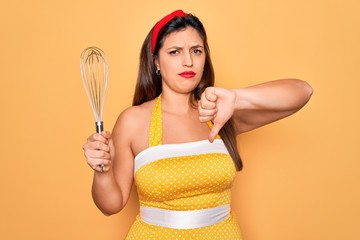 Young hispanic pin up woman wearing fashion sexy 50s style holding cooking whisk blender with angry face, negative sign showing dislike with thumbs down, rejection concept