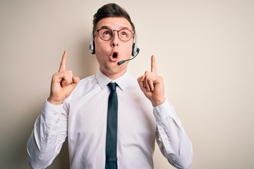 Young handsome caucasian business man wearing call center headset at customer service amazed and surprised looking up and pointing with fingers and raised arms.