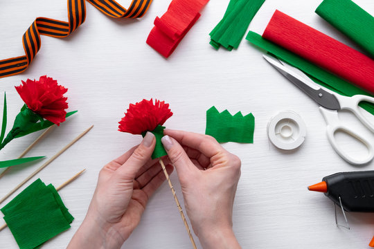 How to make carnation flower at home. Hands making red carnation to Victory Day 9 May. Step by step photo instruction. Step 14. Glue green petals under the bud. Children DIY art project.