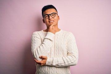 Fototapeta premium Young handsome latin man wearing white casual sweater and glasses over pink background looking confident at the camera with smile with crossed arms and hand raised on chin. Thinking positive.